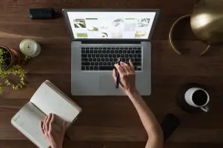 Ilustrasi: Tampak atas tangan seseorang menggunakan laptop dan membuka buku catatan di atas meja kayu.
