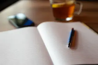 Ilustrasi: Buku catatan terbuka dengan pena di atas meja kayu dekat cangkir minuman.