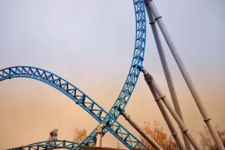 Ilustrasi: Struktur rel roller coaster berwarna biru melengkung dengan latar langit senja.