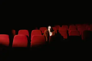 Ilustrasi: Seorang wanita duduk sendirian di antara deretan kursi merah bioskop yang gelap.