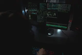 Ilustrasi: Layar monitor komputer menampilkan baris kode di ruangan gelap dengan tangan di atas papan ketik.