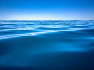Ilustrasi: Permukaan air laut biru bergelombang tenang dengan latar langit biru cerah.