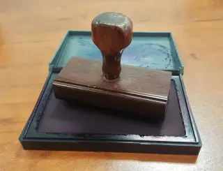 Ilustrasi: Sebuah stempel kayu diletakkan di atas bantalan tinta stempel yang terbuka di atas meja.