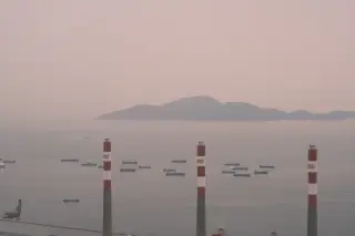 Ilustrasi: Pemandangan cerobong industri dan kapal-kapal di perairan laut di bawah langit berkabut tipis.