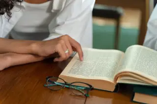 Ilustrasi: Seseorang membaca buku tebal terbuka di meja kayu dengan kacamata di dekatnya.