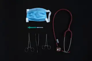 Ilustrasi: Masker medis, jarum suntik, gunting bedah, dan stetoskop tertata rapi di atas latar hitam.