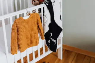 Ilustrasi: Baju rajut anak berwarna kuning tergantung di tepi ranjang bayi putih.