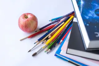 Ilustrasi: Sebuah apel merah, pensil warna, kuas, dan tumpukan buku di atas latar putih.