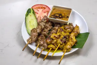 Ilustrasi: Sepiring sate dengan bumbu kacang, irisan timun, dan tomat di atas meja putih.