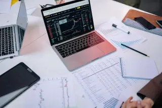 Ilustrasi: Laptop menampilkan grafik pergerakan pasar saham dan dokumen keuangan di atas meja kerja.