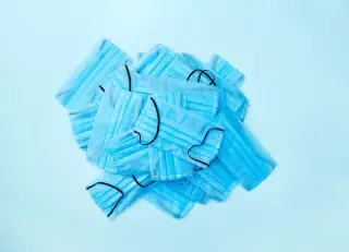 Ilustrasi: Tumpukan masker medis sekali pakai berwarna biru di atas latar belakang biru muda.