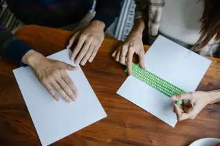 Ilustrasi: Dua orang membaca dan menulis huruf braille di atas kertas putih pada meja kayu.