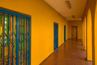 Ilustrasi: Lorong bangunan kosong dengan dinding bercat kuning cerah dan deretan pintu bertralis biru.