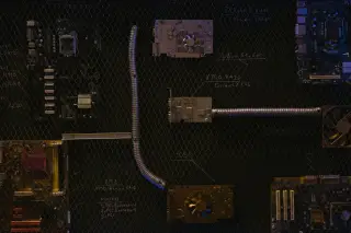 Ilustrasi: Komponen motherboard dan pipa fleksibel tertata di atas latar jaring kawat hitam.