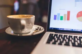 Ilustrasi: Secangkir kopi di atas meja kayu di sebelah laptop yang menampilkan grafik data.