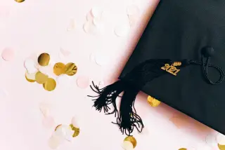Ilustrasi: Topi toga wisuda hitam dengan rumbai bertahun 2021 di atas latar merah muda bertabur konfeti bulat.