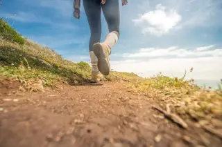 Ilustrasi: Kaki seorang pejalan kaki dengan sepatu trekking melangkah di jalur setapak berbukit.