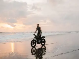 Ilustrasi: Seorang pria mengendarai sepeda motor kustom di tepi pantai berpasir saat senja.