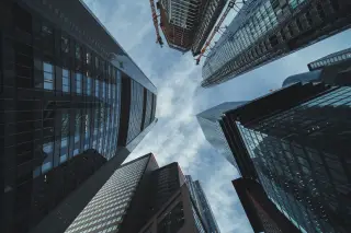Ilustrasi: Sudut pandang rendah menghadap ke atas deretan gedung pencakar langit modern dan langit berawan.