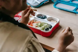 Ilustrasi: Seseorang sedang menyantap makanan dari kotak bekal di atas meja kayu.