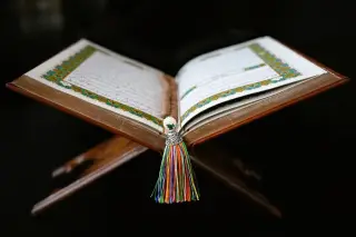 Ilustrasi: Al-Qur'an terbuka di atas rehal kayu dengan tasbih berumbai warna-warni berlatar hitam.
