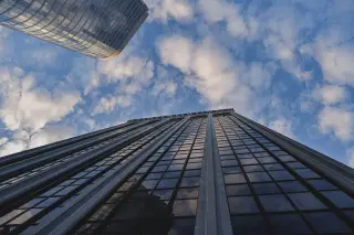 Ilustrasi: Gedung pencakar langit berkaca menghadap ke atas dengan latar langit biru berawan.