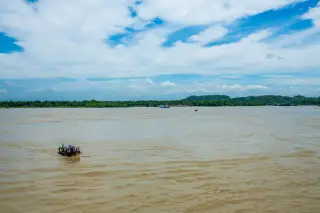 Ilustrasi: Sebuah perahu kecil sarat penumpang melintasi sungai yang sangat lebar di bawah langit berawan.