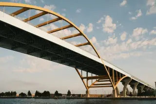 Ilustrasi: Jembatan lengkung berstruktur kuning membentang di atas perairan dengan latar langit biru berawan.