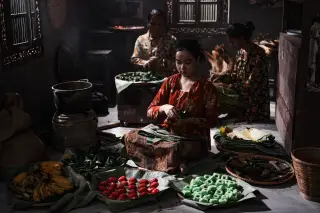 Ilustrasi: Tiga wanita mengenakan kebaya sedang menyiapkan aneka jajanan pasar tradisional di atas lantai.