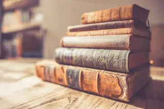 Ilustrasi: Tumpukan buku-buku tebal berjilid kulit klasik di atas permukaan meja kayu.