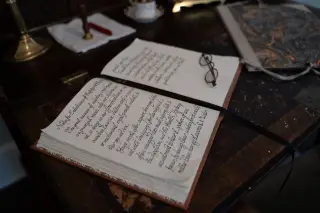 Ilustrasi: Buku catatan terbuka dengan tulisan kaligrafi dan kacamata antik di atas meja kayu.