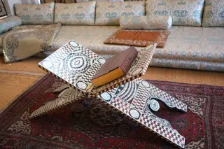 Ilustrasi: Sebuah rehal berukir motif geometris menopang buku tebal di atas karpet bermotif.
