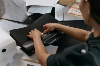 Ilustrasi: Seseorang membuka tas kerja kulit hitam di atas meja kantor yang dipenuhi dokumen.