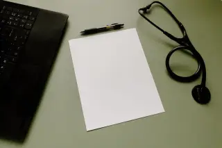 Ilustrasi: Kertas kosong, pena, stetoskop, dan sebagian laptop di atas meja hijau.