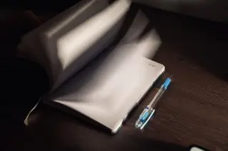 Ilustrasi: Buku catatan terbuka dengan halaman yang membalik dan pulpen di atas meja kayu.