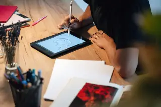 Ilustrasi: Seseorang sedang menggambar karakter menggunakan pena digital di atas tablet di sebuah meja kerja.