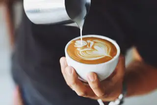 Ilustrasi: Tangan seseorang menuangkan susu dari teko kecil untuk membuat seni latte di cangkir kopi.