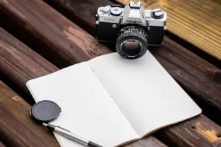 Ilustrasi: Kamera analog, buku catatan terbuka bergaris, dan pena tergeletak di atas meja kayu.