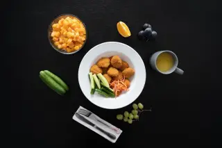 Ilustrasi: Sajian makanan dengan nuget, sayuran, buah-buahan, dan minuman di atas meja hitam.