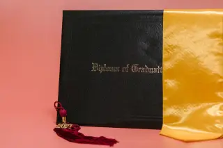 Ilustrasi: Map ijazah hitam bertuliskan Diploma of Graduation dengan rumbai wisuda dan selempang kuning.