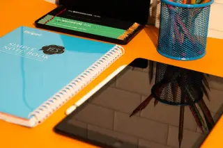 Ilustrasi: Buku catatan, tablet digital, dan perlengkapan pensil di atas meja berwarna oranye.