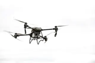 Ilustrasi: Sebuah drone besar dengan baling-baling berputar melayang dengan latar belakang langit putih.