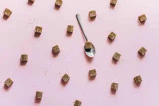 Ilustrasi: Potongan gula kubus cokelat tersebar di atas latar merah muda bersama satu sendok kecil.
