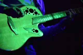 Ilustrasi: Seseorang memainkan gitar akustik dengan sorotan lampu panggung berwarna hijau.