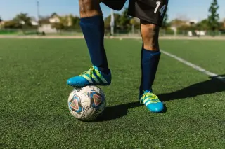 Ilustrasi: Kaki seorang pemain sepak bola mengenakan sepatu biru menginjak bola di atas rumput lapangan hijau.