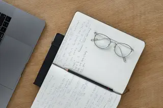 Ilustrasi: Buku catatan terbuka berisi tulisan tangan, kacamata, pensil, dan laptop di atas meja kayu.
