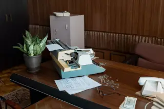 Ilustrasi: Mesin tik antik, kacamata, kertas, dan telepon di atas meja kerja kayu bergaya retro.
