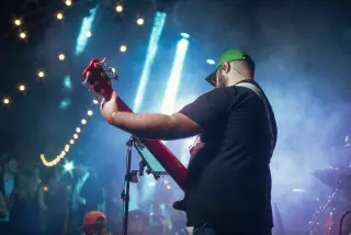 Ilustrasi: Seorang pria tampak dari belakang memainkan gitar bas di panggung konser terbuka dengan sorotan lampu.