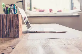 Ilustrasi: Sebuah laptop, alat tulis, dan kertas di atas meja kayu dekat jendela yang terbuka.
