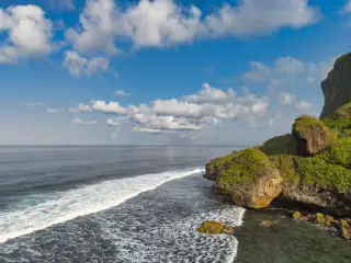 Ilustrasi: Pemandangan tebing karang bervegetasi dan ombak laut di bawah langit biru berawan.